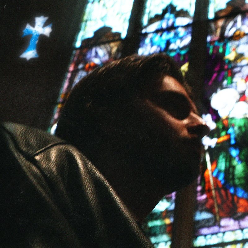Quinn Mills in shadow before a stained-glass window, a cross of light glowing above